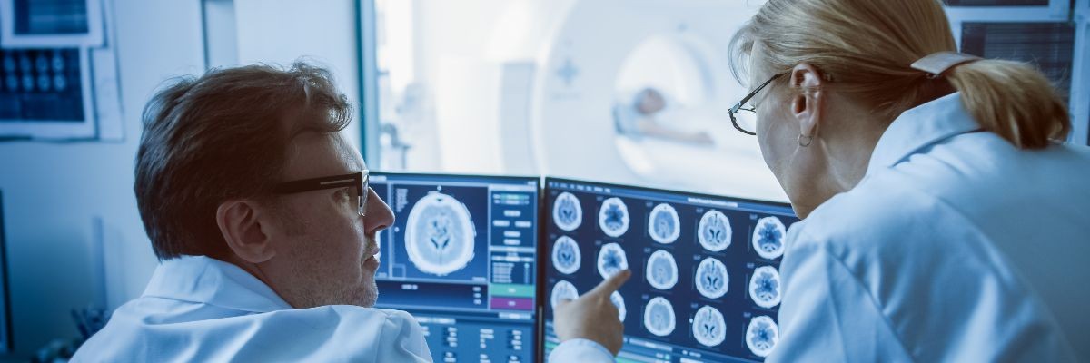 In Control Room Doctor and Radiologist Discuss Diagnosis while Watching Procedure and Monitors Showing Brain Scans Results, In the Background Patient Undergoes MRI or CT Scan Procedure.