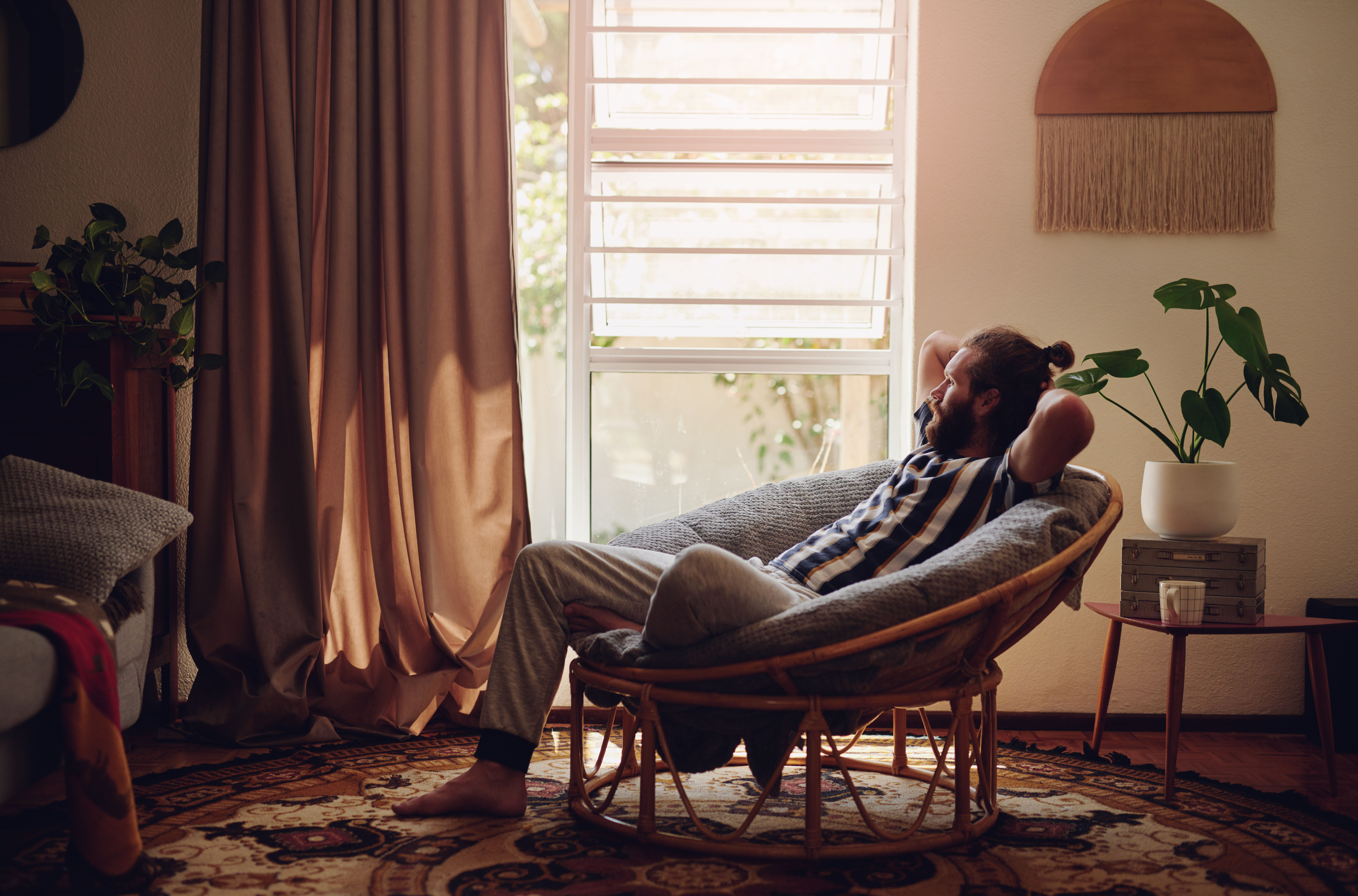 Man with a bun sitting in a bohemian decorated room, looking out of a bright window. . Getty Images photo identifier, 1317367958, Adene Sanchez