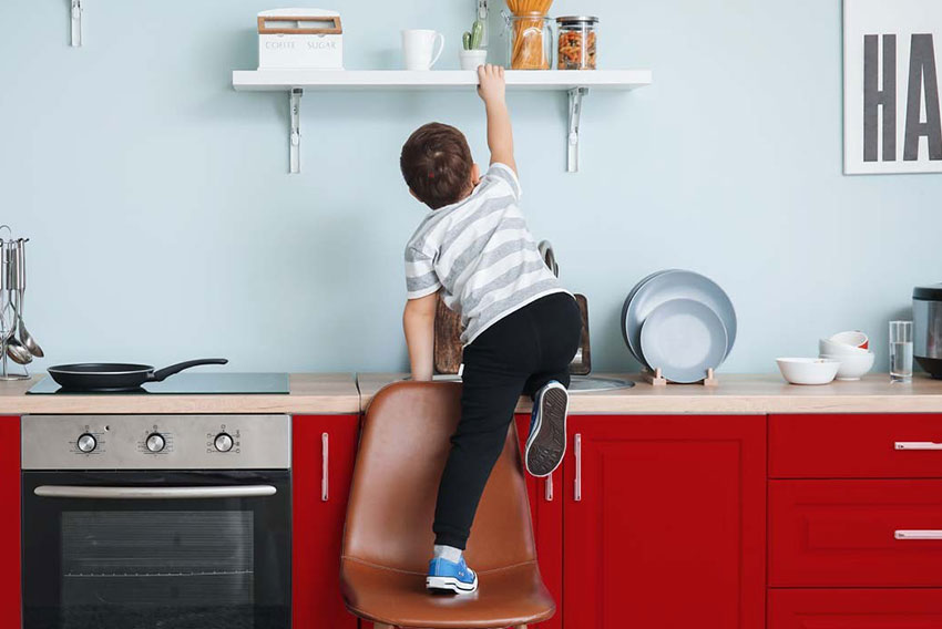 Otrok stoji na stolu v kuhinji in se steguje do police s piškoti. Simbolna slika za zaščito otroka doma.