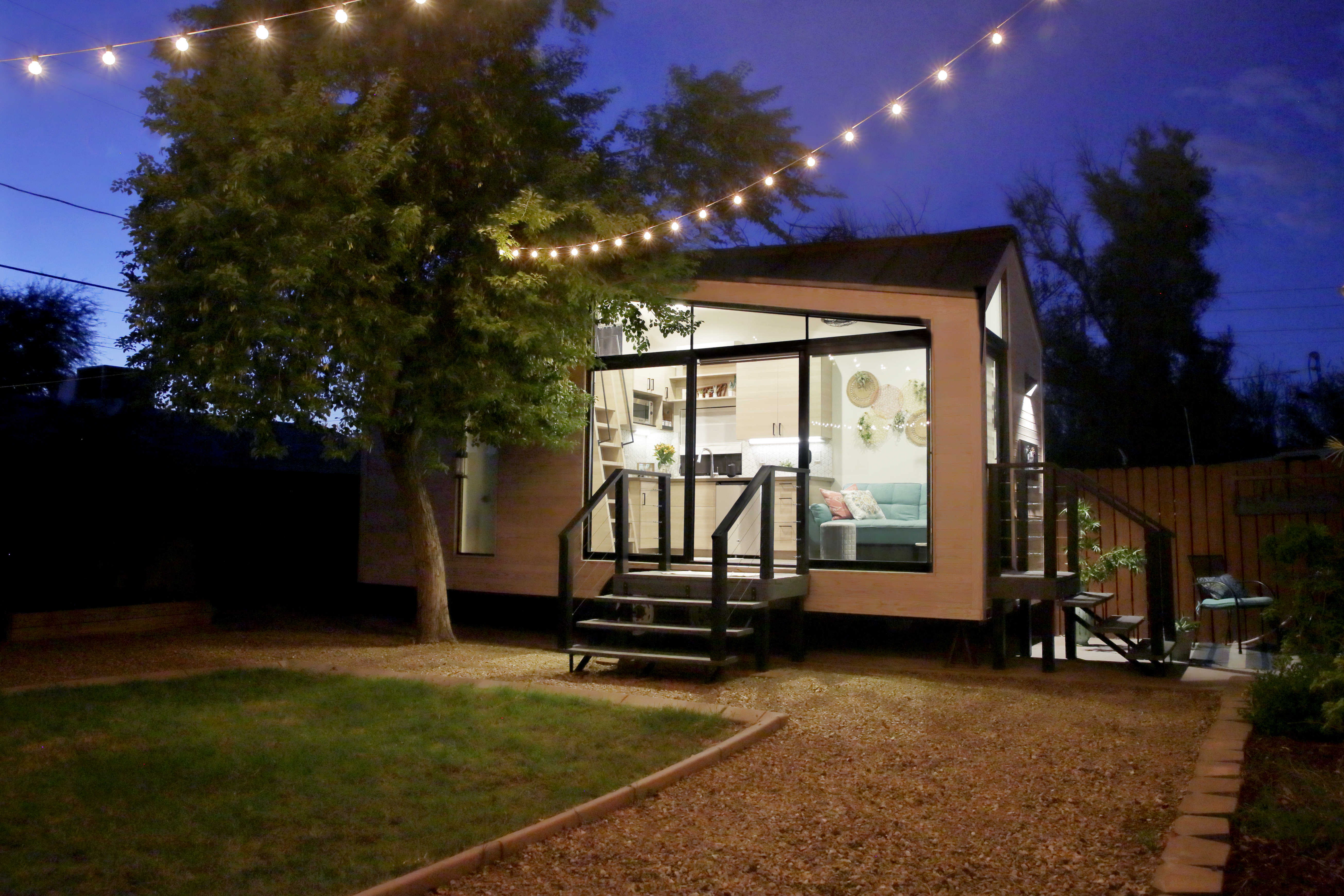 A tiny house with large glass windows, sits in the backyard at night, surrounded by trees and party lights.