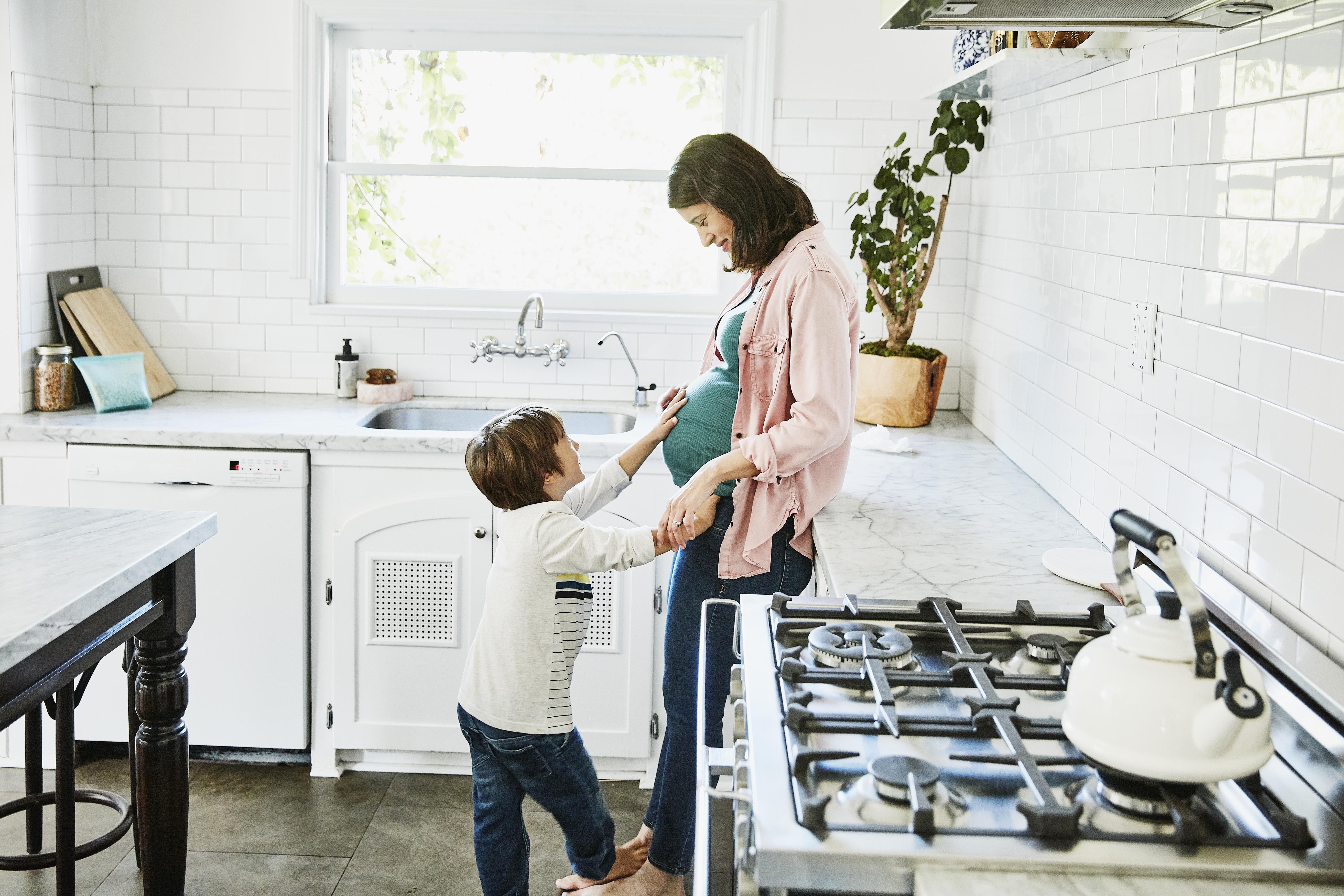 Smiling young son putting hand on pregnant mothers stomach while standing in home kitchen