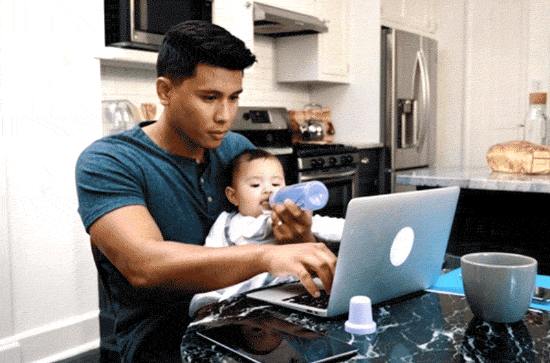 Dad works from home on a laptop while feeding his baby a bottle
