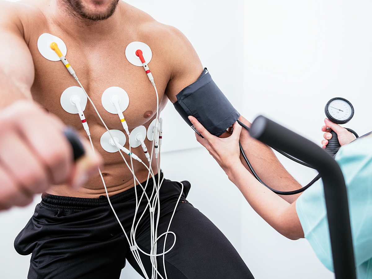 A male patient performs a bicycle exercise test while fitted with multiple electrodes and a blood pressure cuff.
