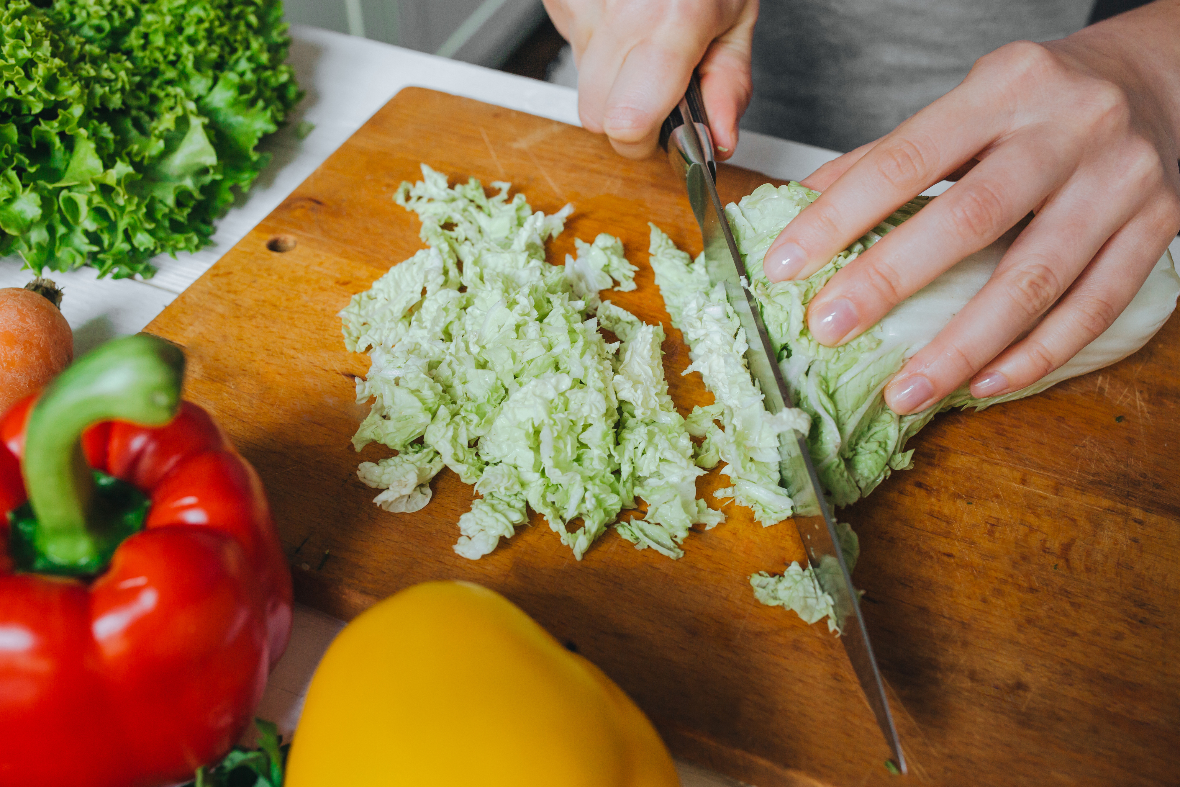 Woman hands cutting fresh green Pekin cabbage on wooden board closeup. White table background. Steel sharp knife reflection.