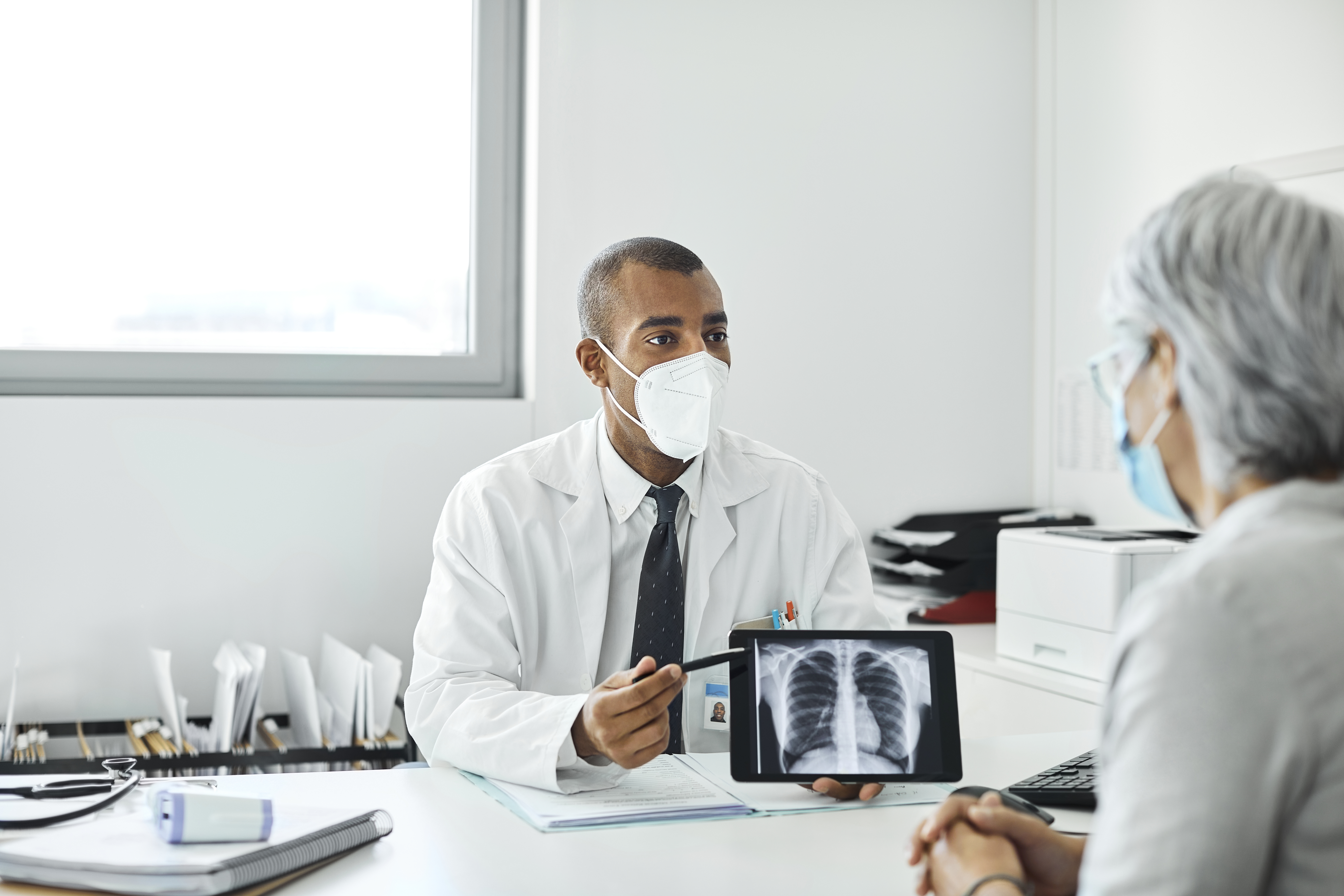 Doctor explaining woman over x-ray on tablet PC. Male medical professional is discussing with patient at clinic. They are wearing protective face masks.