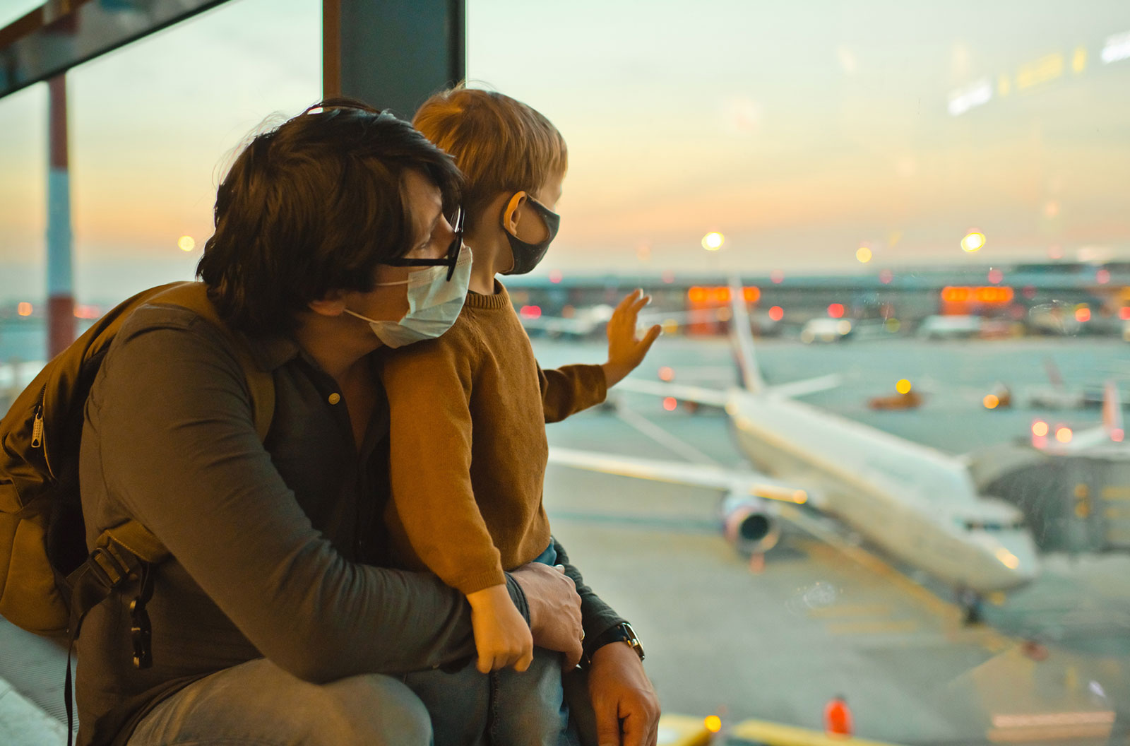 A father and son in masks look outside the window of an airport.