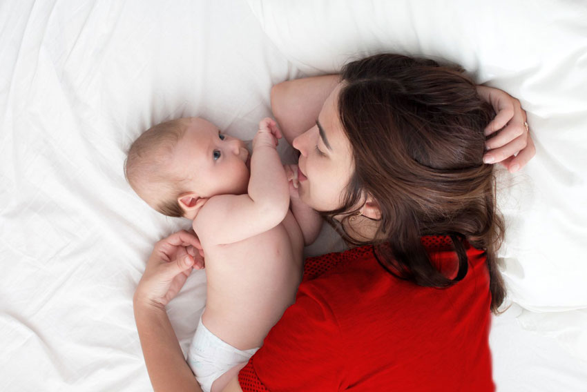 Mati leži na postelji z dojenčkom v plenički in se ga nežno dotika.