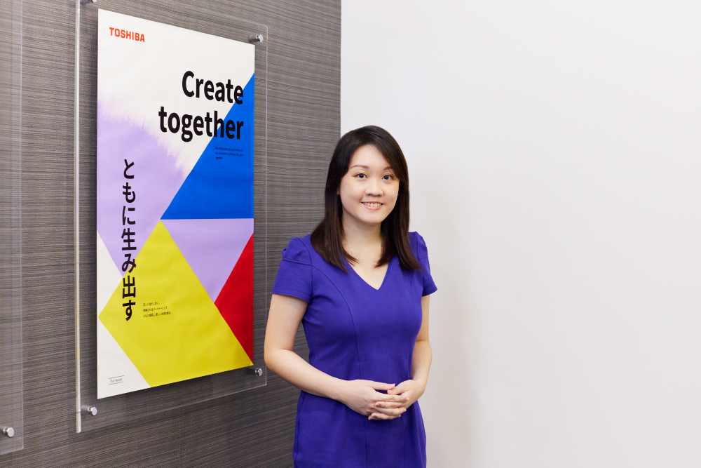東芝グループ理念体系の価値観「ともに生み出す」のポスター前に立つValerie Chin氏