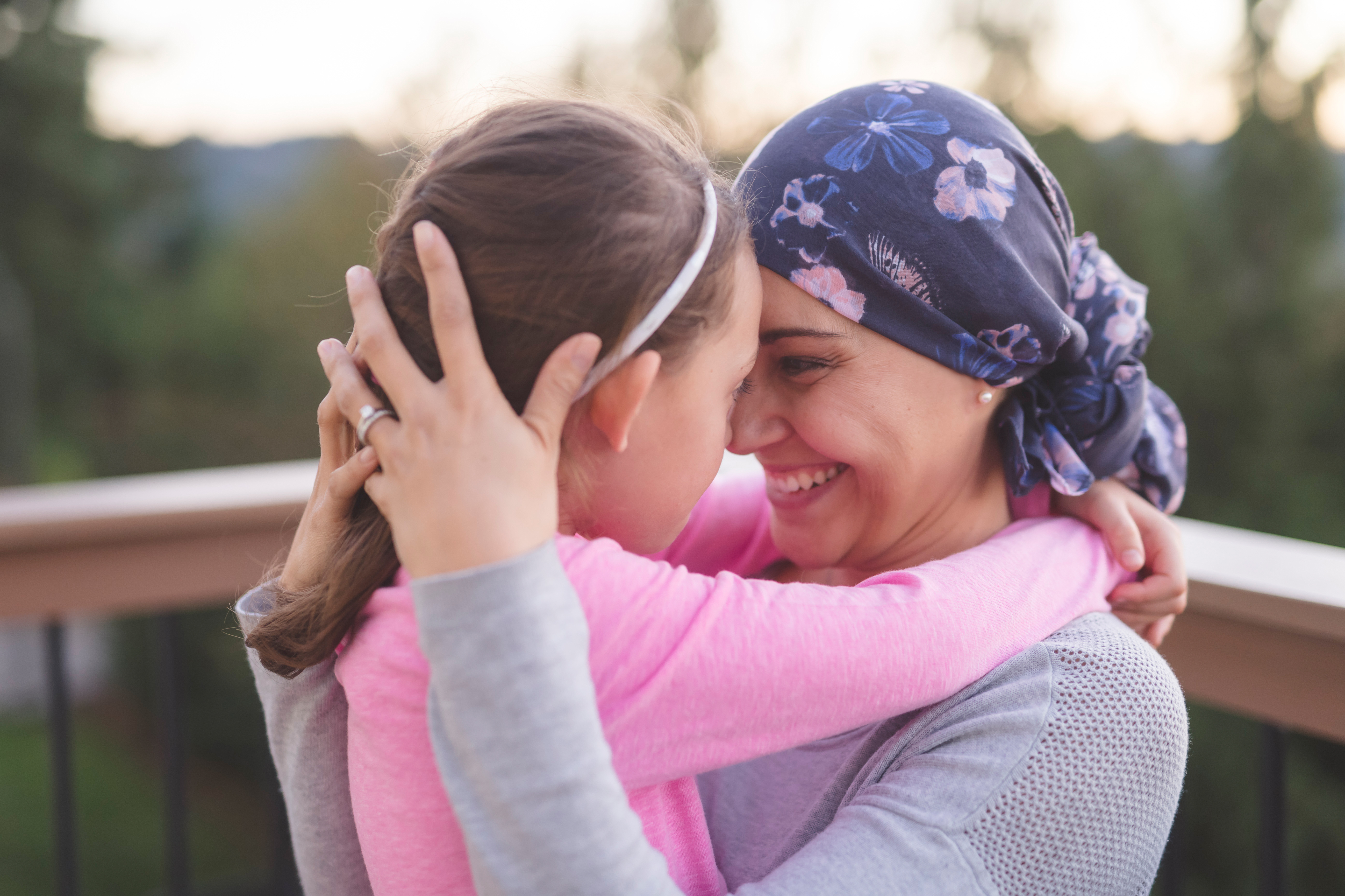 A young mother fighting cancer and wearing a head scarf hugs her daughter and smiles at her deeply as they share a few moments of tranquility together outdoors on a deck. Their faces are pressed together.