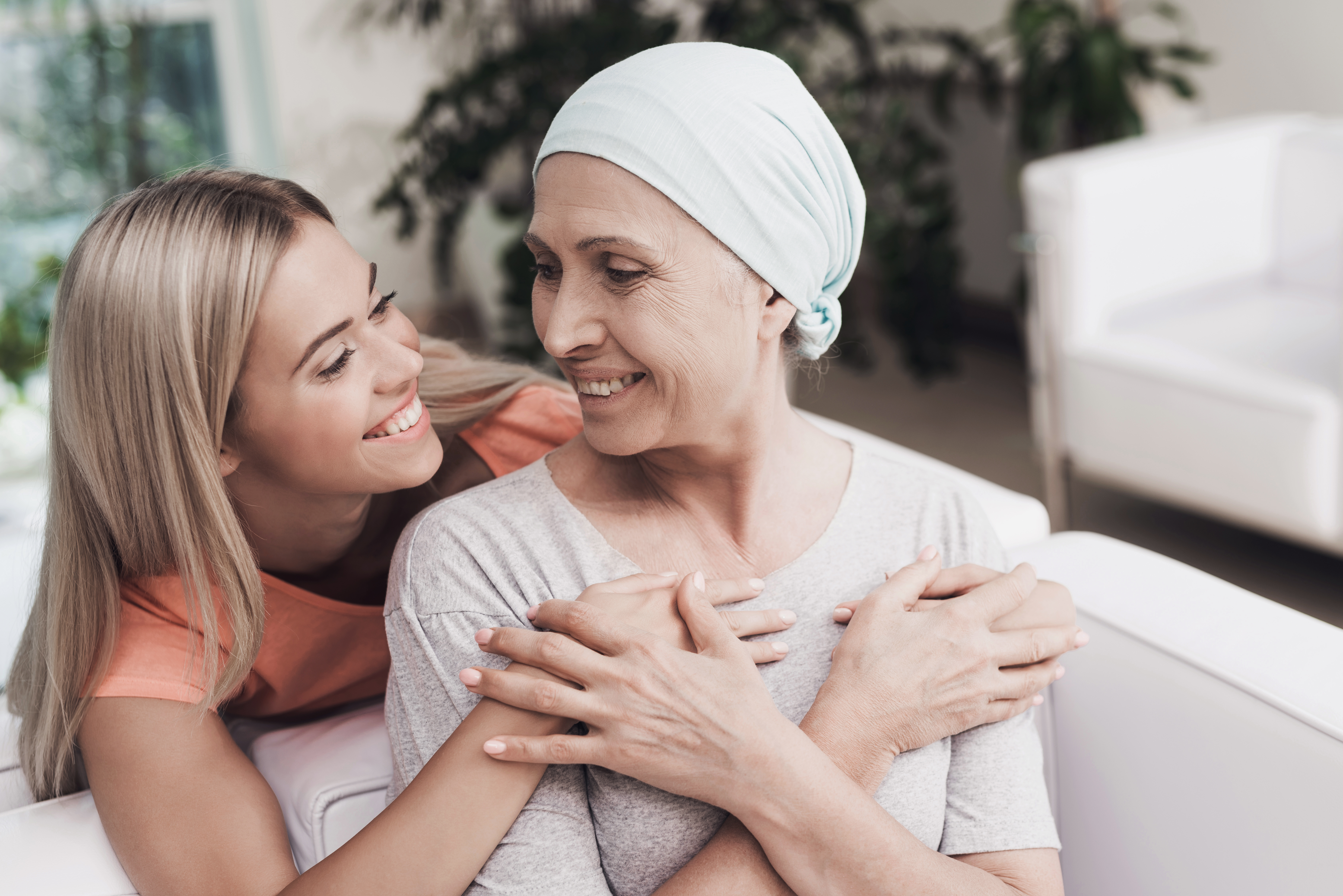 A woman with cancer is sitting on a white sofa next to her daughter. A girl is hugging a woman. They are sitting in the lobby of a modern clinic. Around them flowerpots with flowers.