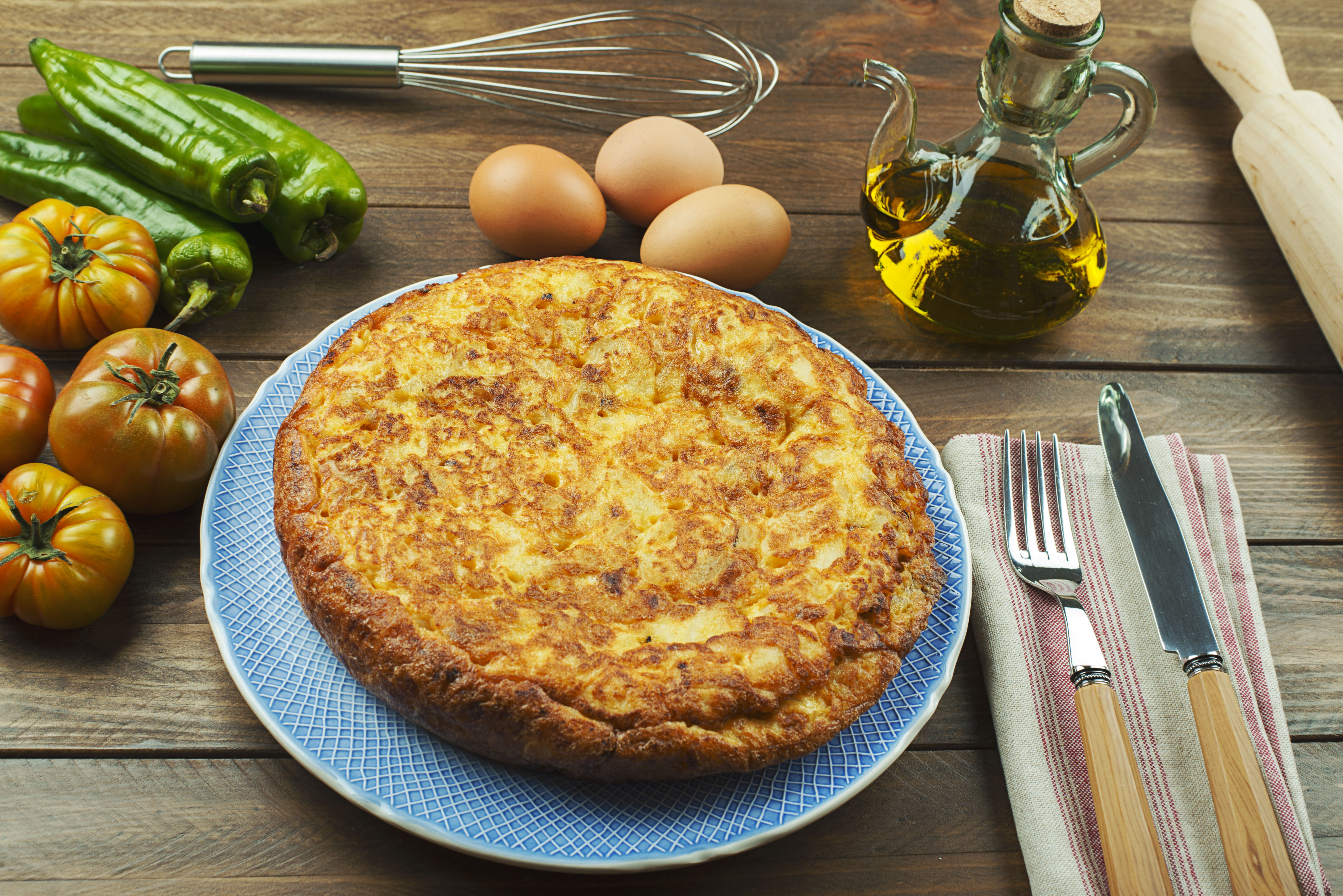 From above plate with typical Spanish omelette, oil, cookware and fresh vegetables on wooden table.