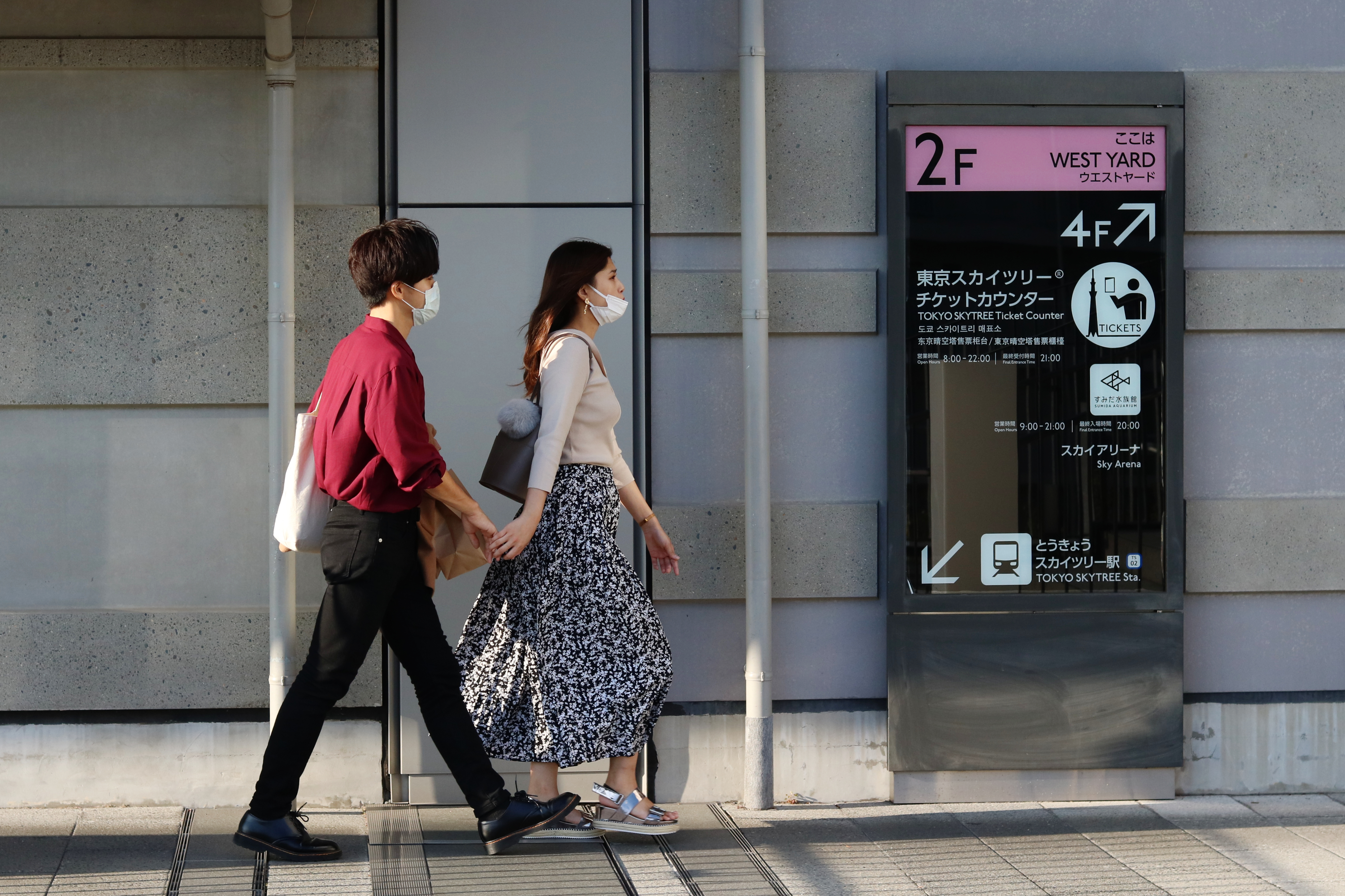 Two people wearing face masks walk past a digital signage solution on a street in the sun.