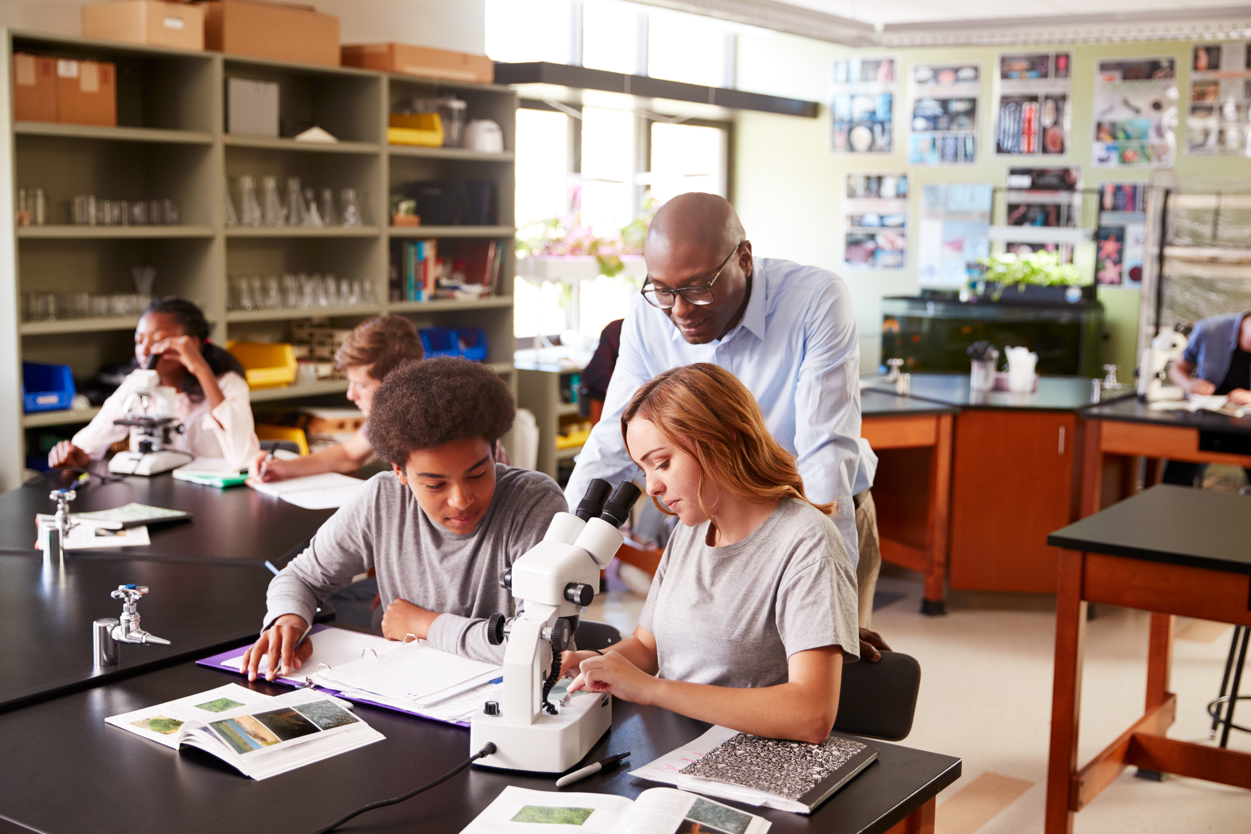 High School Students With Tutor Using Microscope In Biology Class