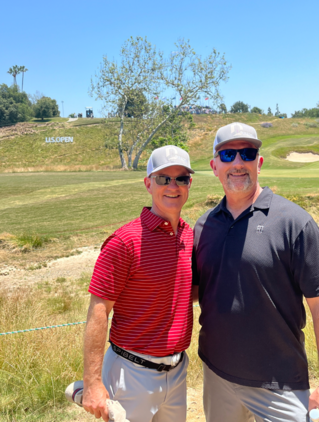 Two men standing together in front of a golf courseDescription automatically generated with medium confidence