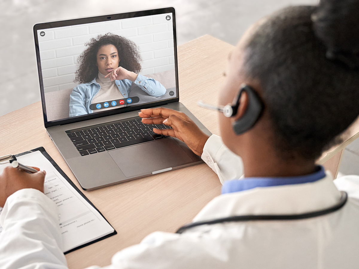 A doctor wearing a headset completes a virtual telemedicine visit with a patient on a laptop. 