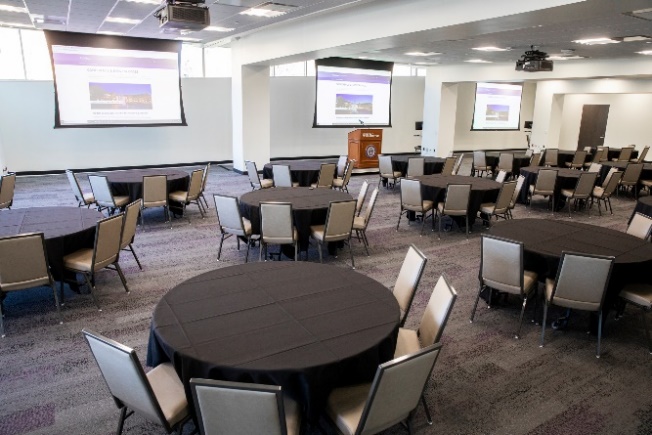 A classroom with circular tables and three laser projectors on screens in the background.