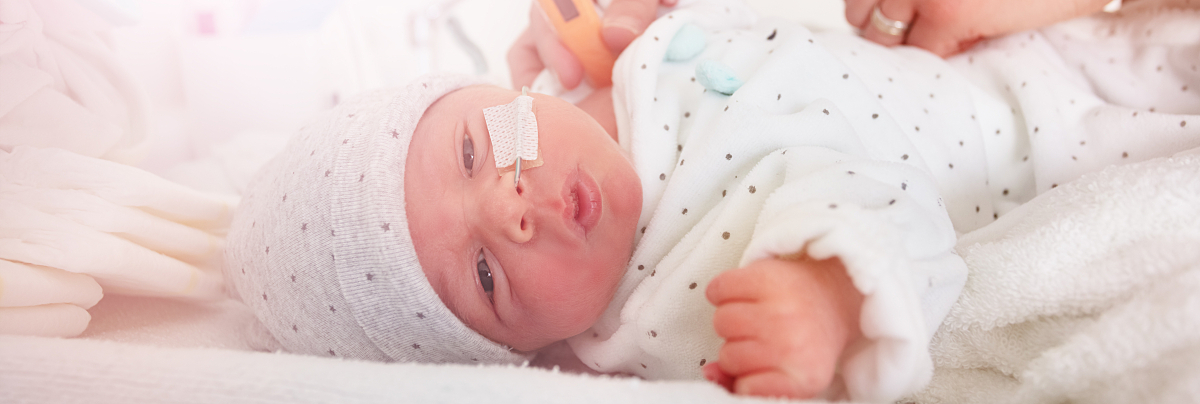 An infant dressed in a bonnet and blanket looks out from the surface of a radiant warmer bed