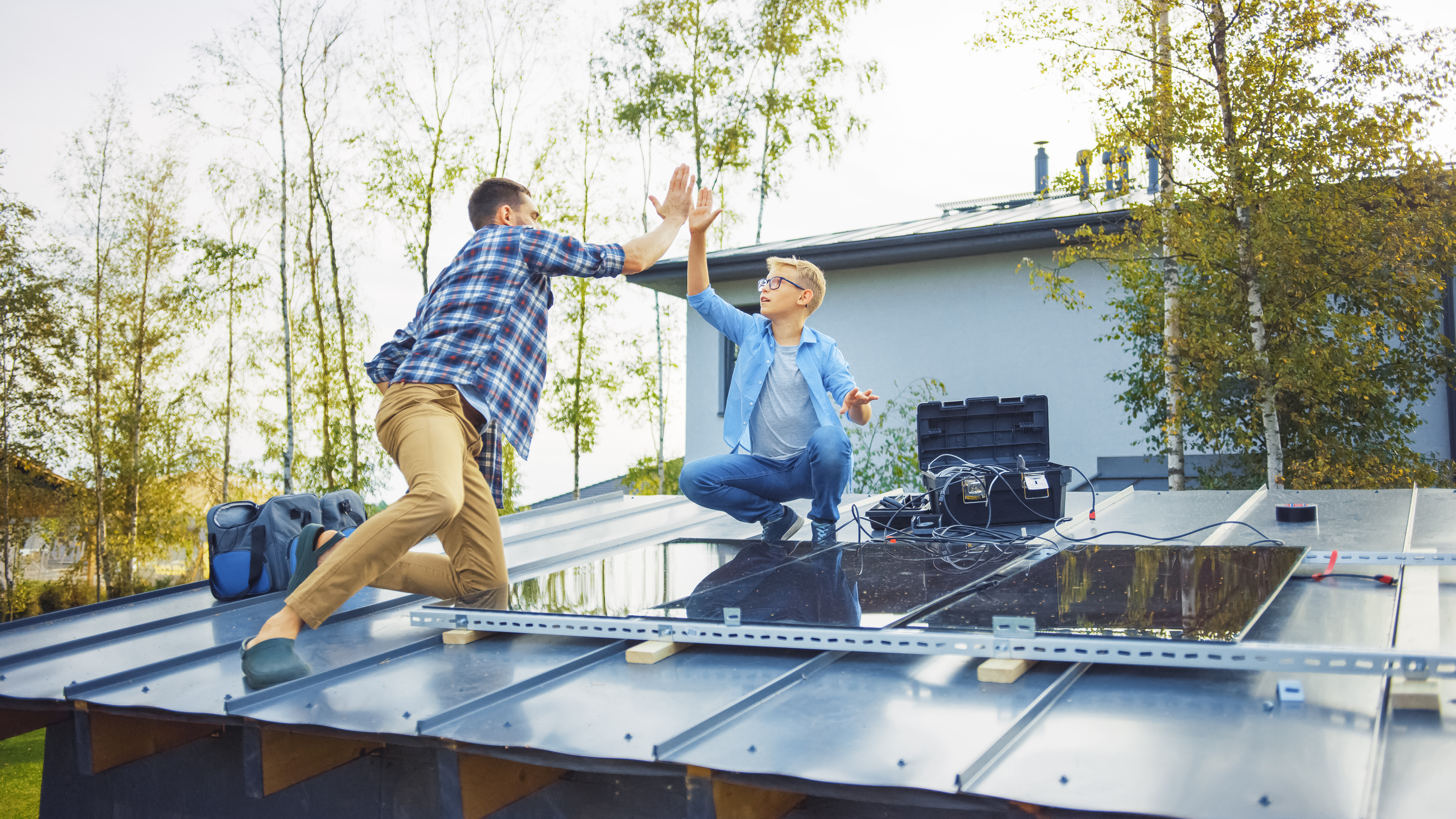 Father and Son Installing Solar Panels to a Metal Basis with a Drill. They Work on a House Roof and Give High Five. Concept of Ecological Renewable Energy at Home and Quality Family Time.