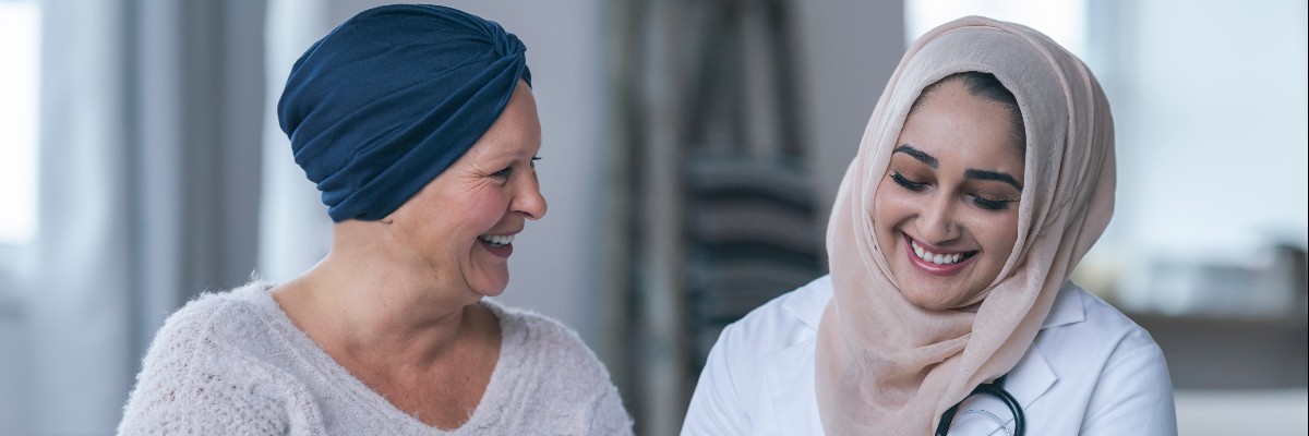 A Muslim female doctor is meeting with a patient. The patient is a mature adult woman with cancer. The patient is wearing a headscarf to hide the hair loss from chemotherapy treatment. The two woman are sitting next to each other on a couch. The doctor is holding a tablet computer. Both women are laughing. They are happy with the patient's recent test results.