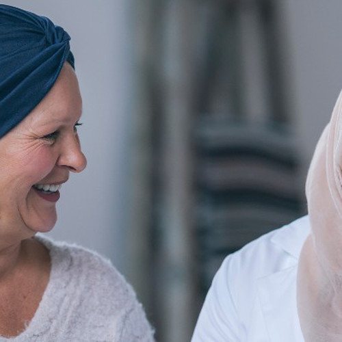 A Muslim female doctor is meeting with a patient. The patient is a mature adult woman with cancer. The patient is wearing a headscarf to hide the hair loss from chemotherapy treatment. The two woman are sitting next to each other on a couch. The doctor is holding a tablet computer. Both women are laughing. They are happy with the patient's recent test results.